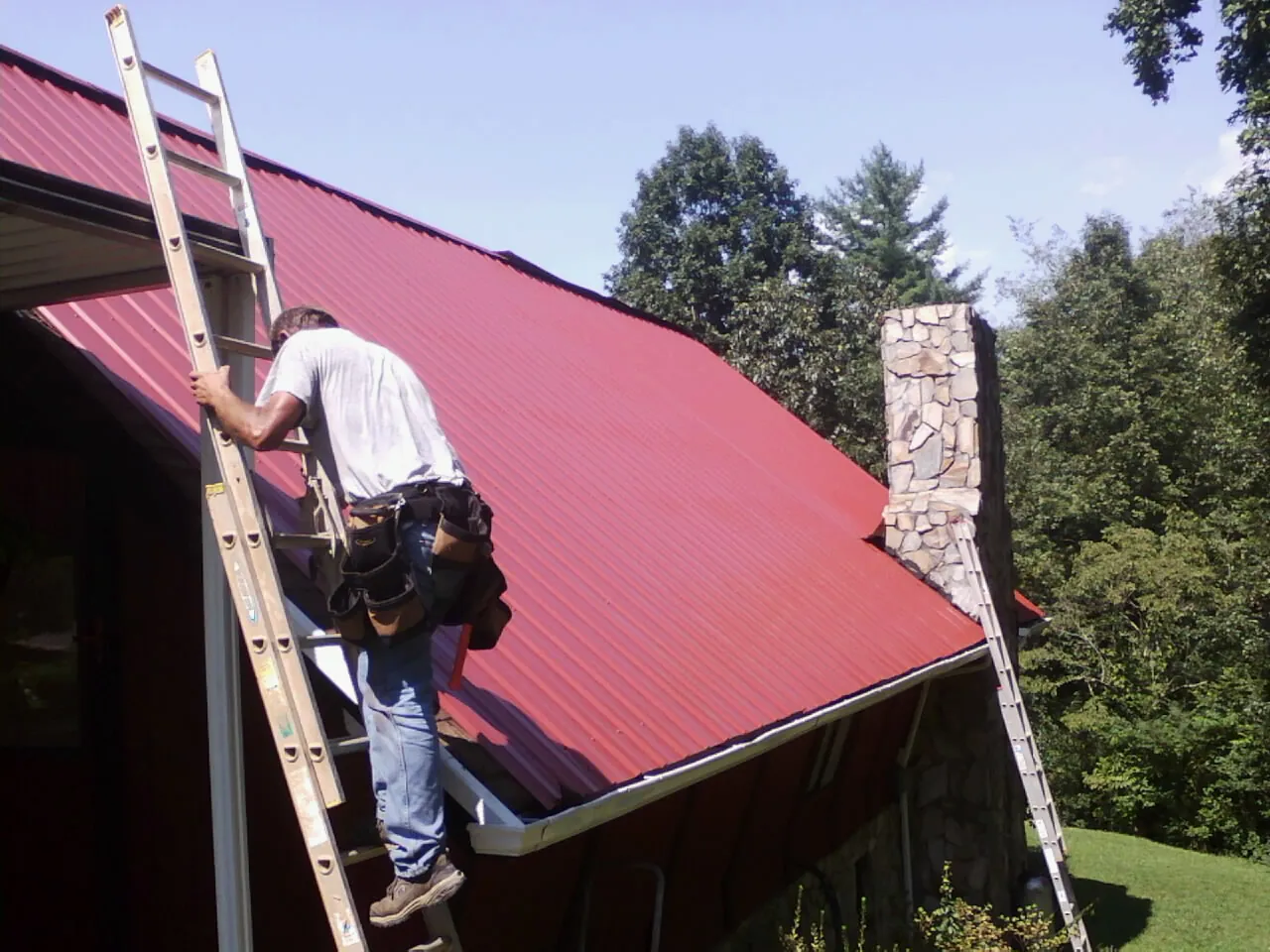 Roofer on a ladder beside a red metal roof and stone chimney — Soffit & Fascia Repair by Roofing Brothers in New York Mills